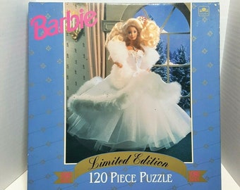 May include: A 120-piece limited edition Barbie jigsaw puzzle featuring a Barbie doll wearing a white gown with a fur trim and a crown. The background is a snowy winter scene.