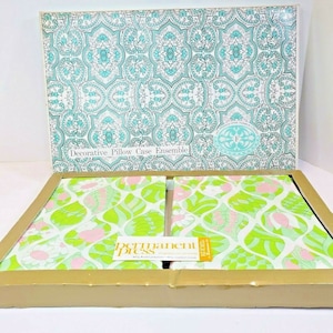 Könnte beinhalten: Ein Set mit zwei dekorativen Kissenbezügen in einer Schachtel. Die Kissenbezüge haben ein grünes und pinkfarbenes Blumenmuster. Die Schachtel ist mit einem türkisfarbenen und weißen geometrischen Muster verziert. Der Text "Decorative Pillow Case Ensemble" und "permanent press" ist auf der Schachtel zu sehen.