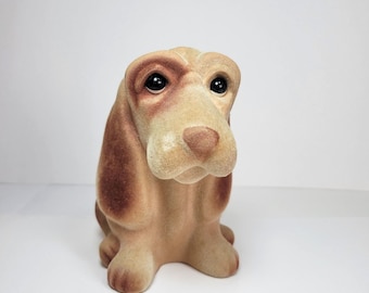 May include: A brown and tan plush basset hound figurine with black eyes. The figurine is sitting with its head tilted down.