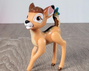 May include: A tan and brown Bambi figurine with a blue butterfly on its tail. The deer has large brown eyes, a white muzzle, and a brown spot pattern on its back. The figurine is made of plastic and stands on four legs.