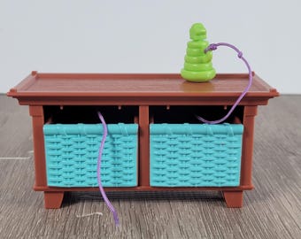May include: A miniature brown wooden cabinet with two turquoise woven baskets. A green, stacked toy and a purple string are on top. The cabinet has a simple design and is set on a wooden surface.