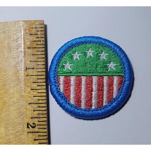 May include: A round patch with a blue border. The top half is green with five white stars, and the bottom half has red and white vertical stripes. The patch is approximately 2 inches in diameter.