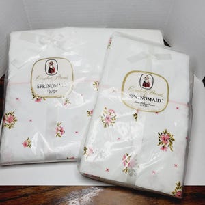 Può includere: Due set di federe bianche in confezione trasparente, legate con fiocchi bianchi. Le federe presentano un motivo floreale con fiori rosa e piccole croci rosse. Ogni confezione ha un'etichetta con la scritta "Combat Percale SPRINGMAID".