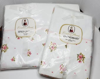 May include: Two white pillowcase sets in clear plastic packaging, tied with white bows. The pillowcases feature a floral pattern with pink flowers and small red crosses. Each package has a label that reads "Combat Percale SPRINGMAID".