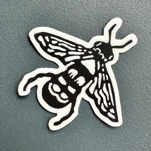 Puede incluir: Pegatina en blanco y negro de una abeja estilizada con las alas extendidas.