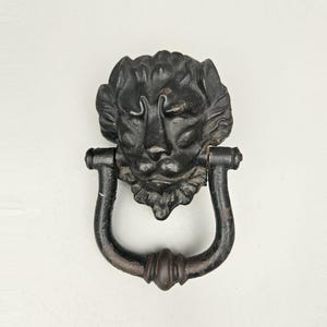 May include: Black cast iron lion head door knocker with a detailed design. The knocker is in the shape of a lion's head with a ring for knocking.