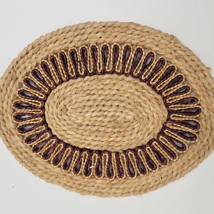 May include: A round, woven, natural fibre coaster with a decorative border of maroon and gold braid.