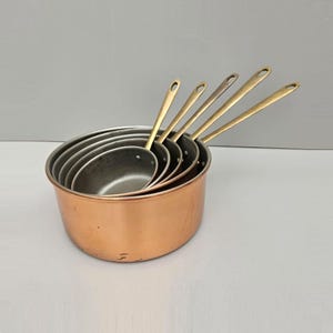 May include: A collection of five copper saucepans with brass handles. The saucepans are stacked inside each other, with the largest at the base. The handles are long and have a loop for hanging. The copper has a warm, reddish-brown colour.