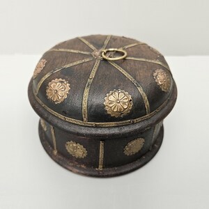 May include: A round wooden box with a domed lid. The lid is decorated with gold metal accents and a small ring handle. The box is also decorated with gold metal accents around the sides.