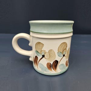 May include: A white ceramic mug with a light blue rim and floral design. The flowers are brown, beige, and light blue.