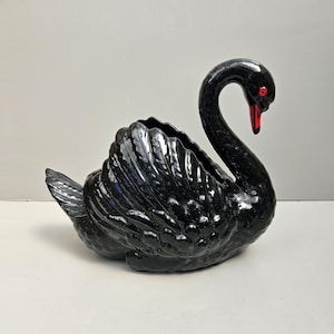 May include: A black swan-shaped planter with a red beak and eyes. The planter features a detailed feather design and a hollowed-out area for plants or decor. The swan is positioned on a white surface.