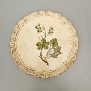 May include: A round, cream-coloured decorative tray with a scalloped edge. It features a painted botanical design of a vine with green and brown leaves. The tray has a distressed, vintage appearance.