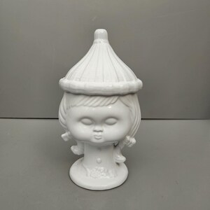 May include: White ceramic figurine of a girl with a conical hat. The figurine has a simple, stylized design and is a decorative object.