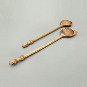May include: Two wooden spoons with long handles and rounded bowls. The spoons are made of light-coloured wood and have decorative, carved handles. One spoon has a round bowl, and the other has a square bowl.