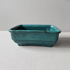 May include: A turquoise ceramic planter, rectangular in shape with a glossy finish. The planter has a raised rim and rests on four small feet. The sides are detailed with horizontal ridges. Suitable for small plants, it measures approximately 20 cm in length.