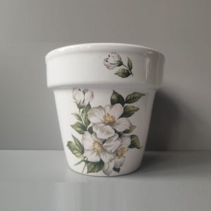 May include: A white ceramic flower pot featuring a floral pattern of white blossoms with yellow centres and green leaves. The design includes a cluster of flowers on the main body and a single small bud near the top rim of the tapered vessel.