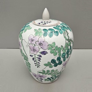 May include: A white ceramic jar with a lid, decorated with a floral pattern of purple and green flowers and leaves.