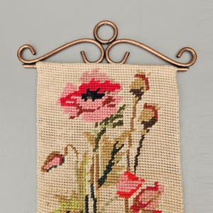 May include: A vintage needlepoint tapestry featuring a floral design of red and pink poppies with green leaves and stems. The tapestry is mounted on a decorative copper-coloured metal hanger. The background is a light beige colour.