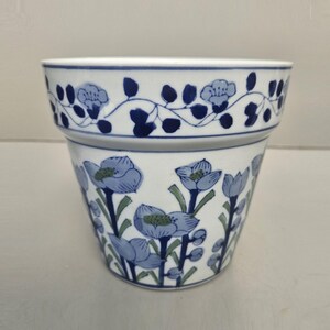 May include: White ceramic plant pot with a blue floral design. The pot has a band of blue flowers and leaves around the rim, with larger blue flowers and green stems decorating the body. A decorative piece for plants.