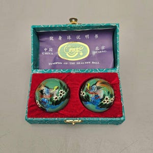 May include: Two green health balls with a painted bird and floral design, presented in a teal box with a red velvet interior. The box contains a paper insert with Chinese text and the words "Synopsis of the Healthy Ball."