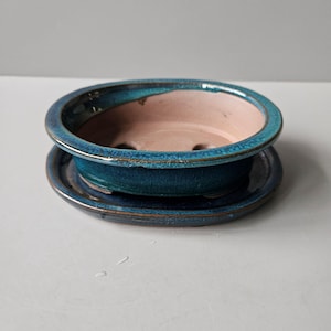 May include: A teal ceramic bonsai pot with a matching saucer. The pot has a round shape with a wide rim and drainage holes. The interior is a light pink colour, contrasting with the teal exterior. The saucer is a smaller, matching teal dish.