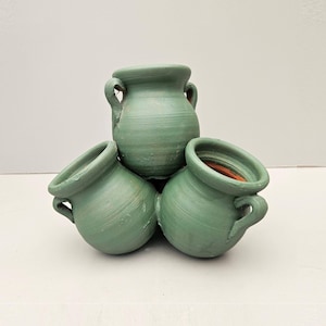 May include: A cluster of three sage green ceramic pots arranged in a pyramid formation. Each pot features a rounded body, a wide circular opening, and two small side handles. The exterior surface has a matte finish with visible horizontal texture.