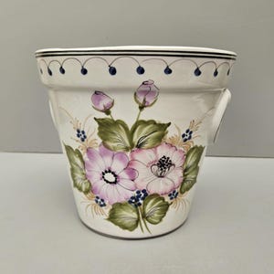 May include: Cream ceramic plant pot with a hand-painted floral design. The pot features pink and purple flowers, green leaves, and blue accents. The rim has a black and blue dotted pattern. Includes two small handles.