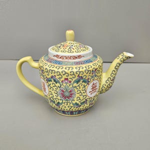 May include: A yellow teapot with a floral design and Chinese characters. The teapot has a lid and a spout. The design is in shades of blue, red, and green.