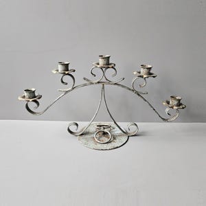 May include: A vintage-style metal candelabra in off-white, featuring five candle holders. The candelabra has a curved, arched design with decorative scrollwork and a circular base with a ring. The candle holders are small and show signs of age.