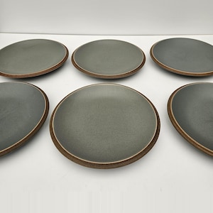 May include: Six grey ceramic plates with a speckled finish and a brown rim are arranged on a white surface. The plates are round with a slightly irregular shape, suggesting a handmade quality. The plates are suitable for serving food.
