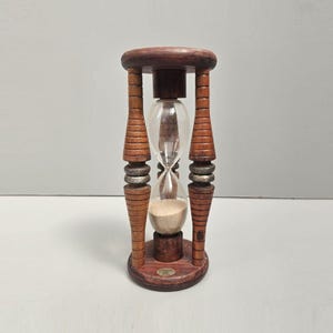 May include: A vintage hourglass with a wooden frame and clear glass bulb. The hourglass has a dark brown finish with decorative silver bands and contains beige sand. The hourglass is a decorative item.
