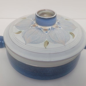 May include: A white ceramic casserole dish with a blue lid and floral design. The dish has a blue and white striped pattern on the bottom.