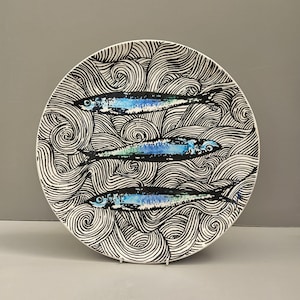 May include: A circular ceramic plate featuring a black and white wave design. Three fish, coloured blue and black, are painted on the plate. The plate is displayed on a white metal stand against a grey background.