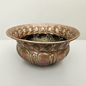 May include: A copper bowl with an embossed design. The bowl has a wide rim and a rounded body. The embossed pattern includes oval shapes and curved lines. The interior is dark, and the exterior shows signs of age, with some green oxidation.