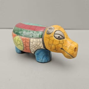 May include: A ceramic hippo figurine with a patchwork design. The hippo's body features sections of red, green, and yellow, while the head is yellow. The legs are blue, and the figurine has a crackle glaze.