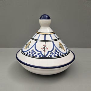 May include: A white ceramic tagine pot with a conical lid. The lid is decorated with blue, yellow, and pink floral patterns and a blue knob. The base has a blue rim. This traditional cookware is used for slow-cooked stews.