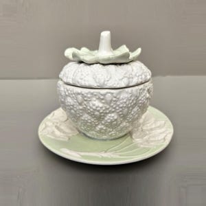 May include: A white ceramic lidded jar with a textured, bumpy surface resembling a fruit, topped with a pale green leaf-shaped handle. The jar sits on a matching round saucer featuring a pale green leaf design embossed on the white ceramic surface.