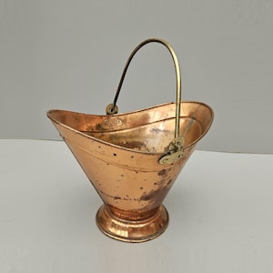 May include: A polished copper coal scuttle with a wide, flared opening and a rounded base. The vessel features a curved brass handle attached with circular rivets on each side. The metal surface shows natural patina and minor dark spots throughout.