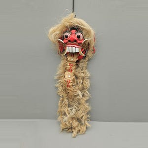 May include: A red and white mask with a fierce expression, featuring large white teeth and a straw-like mane. The mask has a long, segmented neck with red accents and is suspended by a black string. The overall design suggests a cultural or ceremonial artifact.