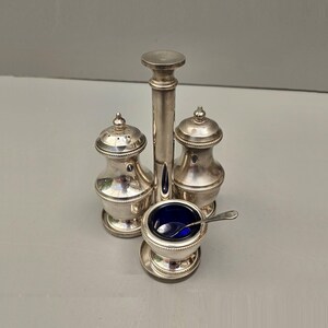 May include: A silver condiment set comprising two shakers, a small bowl with a blue interior, and a central handle. A tiny spoon rests in the bowl. The set is made of silver-coloured metal and sits on a light grey surface.
