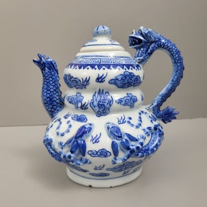 May include: A blue and white porcelain teapot with a dragon handle and dragon details on the body. The teapot has a bulbous body with three tiers and a lid with a geometric design.