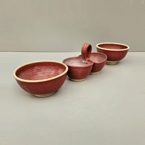 May include: A collection of four handmade ceramic bowls in a deep red colour with a cream-coloured rim. The set includes two round bowls, a double bowl with a handle, and a smaller bowl. The bowls are arranged on a light grey surface.