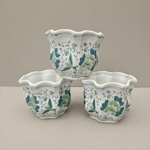 May include: Three white ceramic plant pots with a scalloped rim. Each pot is decorated with a blue and green floral pattern. The pots are stacked on top of each other.