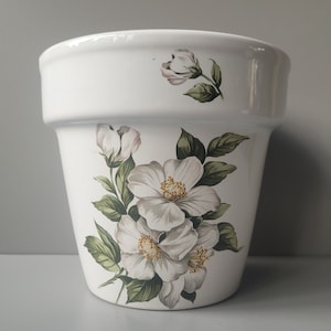 May include: White ceramic flower pot with a floral design featuring white flowers and green leaves.