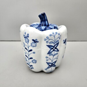 May include: A white ceramic pepper shaker with a blue floral design and a blue lid shaped like a pepper.