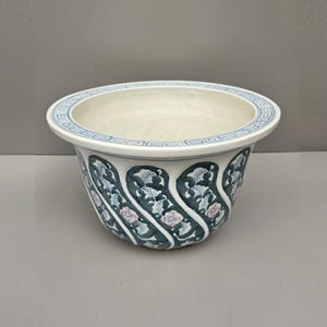 May include: A ceramic planter featuring a wide, flared rim with a blue geometric Greek key pattern. The exterior body is decorated with repeating diagonal bands of dark teal foliage and small pink flowers against a white background.