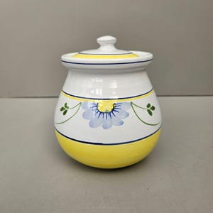 May include: A white ceramic jar with a lid, featuring a yellow base and rim, and a blue floral design. The jar has a rounded shape and is decorated with green vine-like accents. The lid has a small knob.