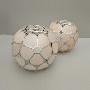May include: Two spherical lampshades with a floral design. The lampshades have light pink petals and silver-coloured metal frames. Each lampshade has a round opening at the top for a light fitting. The background is a plain light grey.