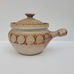 May include: A white ceramic casserole dish with a brown lid and handle. The dish has a decorative pattern of brown circles around the rim.