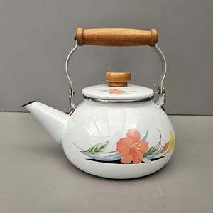 May include: White enamel teapot with a floral design featuring a pink flower and green leaves. The teapot has a wooden handle and a wooden knob on the lid.